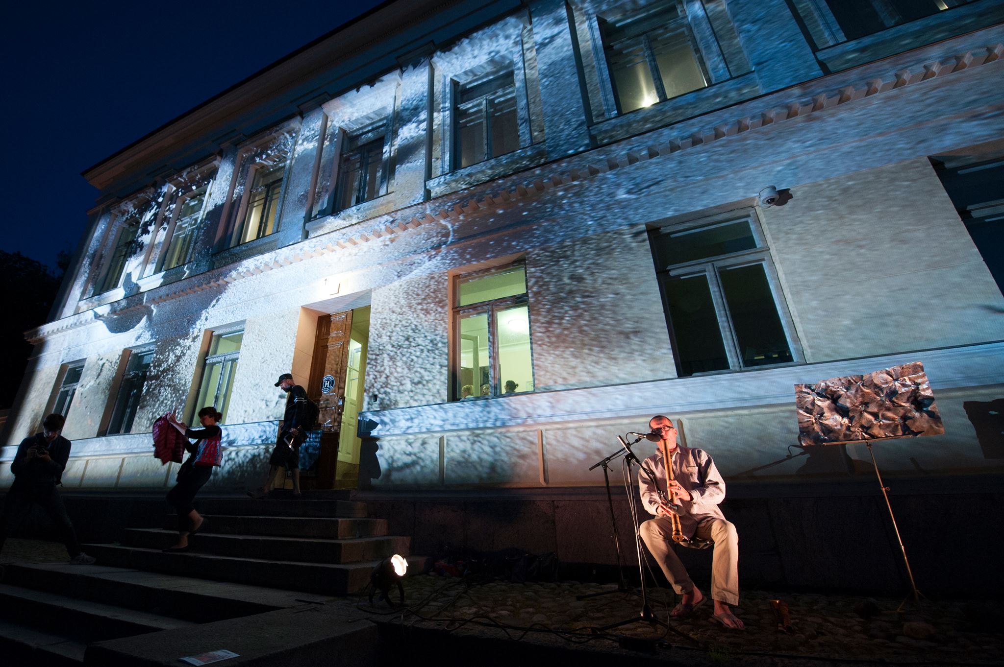 Tales from Frontiers blue facade projection Jone Takamäki playing flute on the foreground